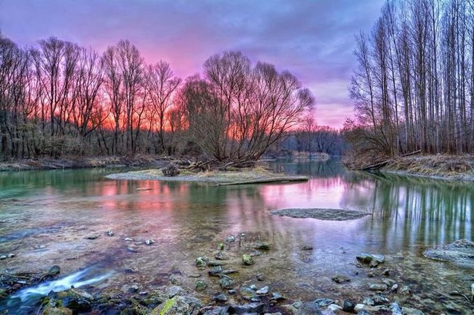 A serene landscape photograph showing a river at sunset or sunrise, with pink and purple hues reflecting in the calm water, surrounded by bare winter trees and rocky shores.