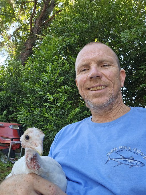A smiling older man wearing a blue 'Reed Hill Cove' t-shirt holding a white turkey, with green foliage and a red lawnmower visible in the background.