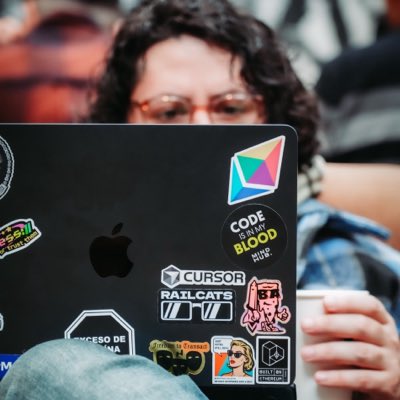 A person with curly hair works on a laptop covered in stickers, including 'CODE IS NOT BLOOD RIP', 'CURSOR', and 'RAILCATS'.