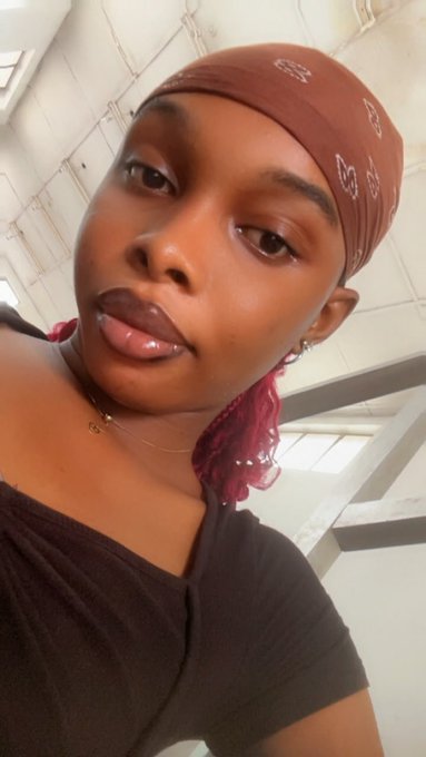 A close-up selfie of a young woman wearing a brown bandana, a black top, a small necklace, and lip gloss, with pink hair extensions visible.