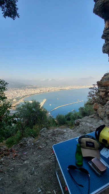 A peaceful weekend view from a rocky hillside overlooks a sprawling coastal city and harbor, with a blue mat, green bottle, sunglasses, phone, and other gear in the foreground. The post’s comment about resting from crypto and finding freedom in forex is reinforced by the serene, off-the-clock scene, while the cigarette butts remain a visible eyesore.