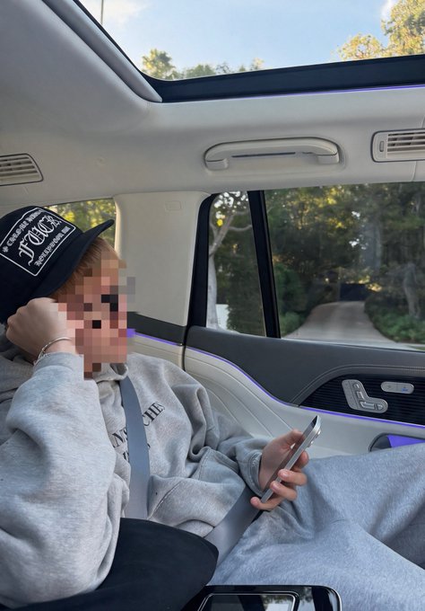 A person wearing a black "Fancy" cap and gray hoodie sits in the back of a luxury car with a sunroof, using a smartphone with their face pixelated for privacy.
