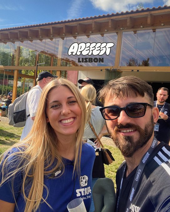 A selfie of a smiling woman in a blue staff shirt and a man wearing sunglasses with a 'CRYPTO' lanyard, taken at the APFEST LISBON event outside a building with a covered patio.