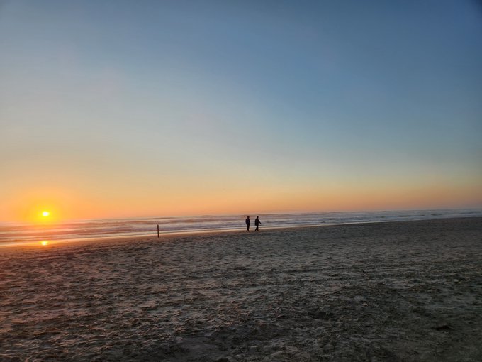 Two silhouettes of people walk along a sandy beach at sunset, with the sun low on the horizon casting an orange glow across the water.