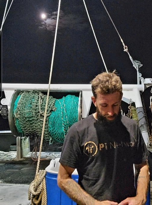 A bearded man wearing a dark grey t-shirt with the logo "PILISANMORA" stands at night on what appears to be a boat, with large spools of green fishing net behind him.
