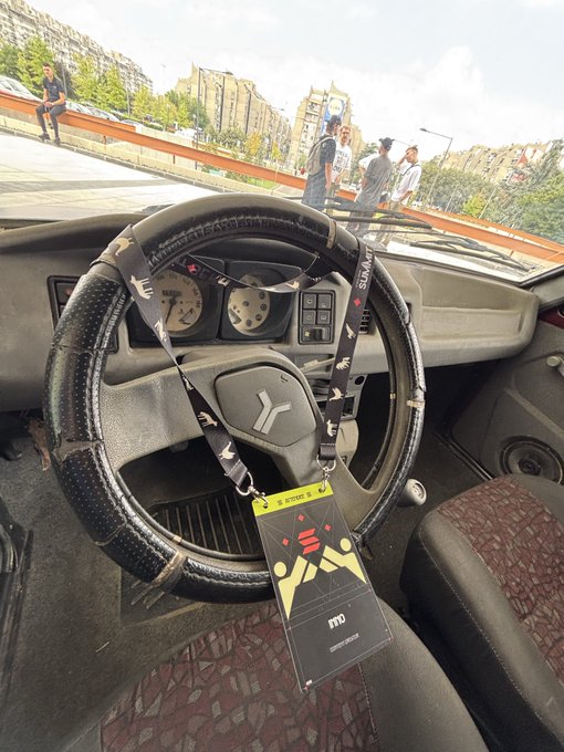 An interior view of a car featuring a worn black steering wheel with a 'BYD' lanyard hanging from it, showing a street scene with pedestrians and apartment buildings visible through the windshield.