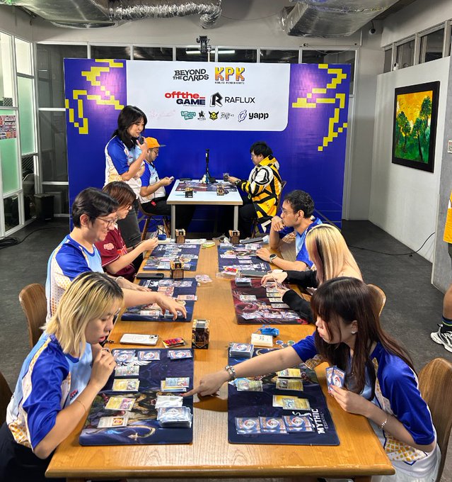 A group of people in blue and yellow jerseys play a card game at a tournament with banners for 'BEYOND THE CARDS' and 'KPK'.
