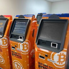 A row of orange Bitcoin ATMs branded with the text "BUY AND SELL BITCOIN" and the iconic ₿ logo are lined up indoors, likely illustrating the type of kiosks affected by Albuquerque's new ordinance banning such crypto transaction machines.