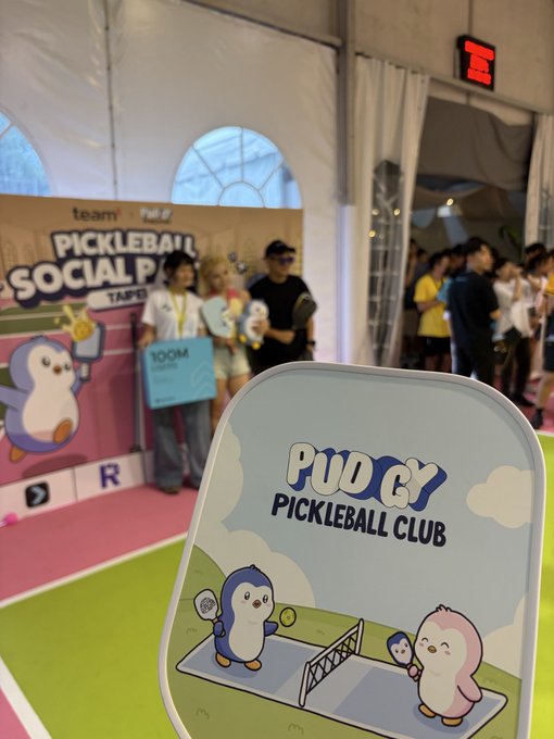 An event space showing a "PUDGY PICKLEBALL CLUB" sign with cartoon penguin mascots, with people gathered in the background near a "PICKLEBALL SOCIAL P" banner.