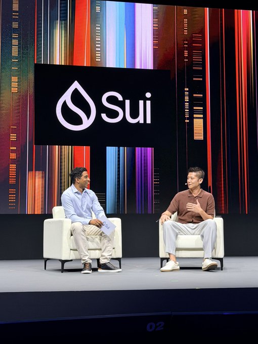 Two men sit in white chairs on a stage in front of a large screen displaying the 'Sui' logo.