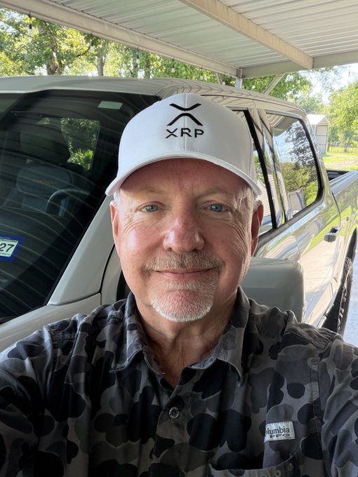 A confident man wearing a white "XRP" cap and a Columbia camo shirt poses for a selfie in front of his pickup truck, channeling crypto optimism. His bold, forward-looking gaze matches the rallying spirit of "Take on the day!"