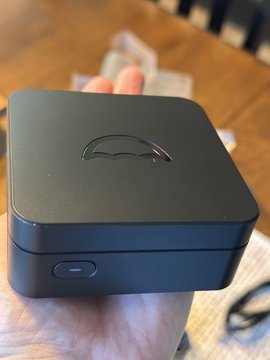 A hand holds a small, black square electronic device with a car logo on top, likely a base station or hub, with a small button on the side.
