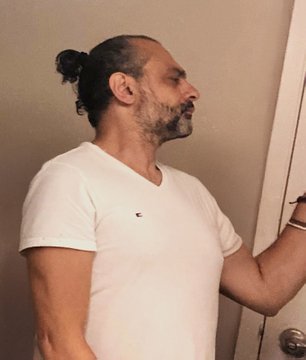 A side-profile shot of a man with dark hair tied in a bun, a beard, and a white V-neck t-shirt, standing near a white door.