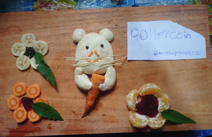A creative food art display on a wooden board featuring a mouse-shaped bun holding a carrot, surrounded by fruit and vegetable flower arrangements, with a handwritten note reading "RollerCoin @hayshop7926432".
