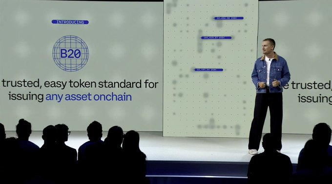 The image shows a man standing at a podium with a large screen behind him displaying information about a new cryptocurrency called Coinbase. The man appears to be giving a speech or presentation, possibly discussing the features of this new digital currency. There are several people in the audience, indicating that it could be an event or gathering related to cryptocurrency and blockchain technology.