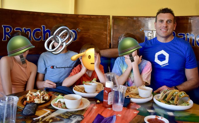 The image shows a family sitting around a dining table at a restaurant or diner, enjoying their meal together. There are four people present - two adults and two children. The man is wearing a blue shirt with "chainlink" written on it, while the other person has a green helmet on. They all have smiles on their faces as they share food and conversation.

The table is set with various dishes such as bowls of soup, plates of food, cups, forks, knives, spoons, and bottles. The scene captures a moment of togetherness and bonding for the family members during their mealtime at the restaurant or diner.