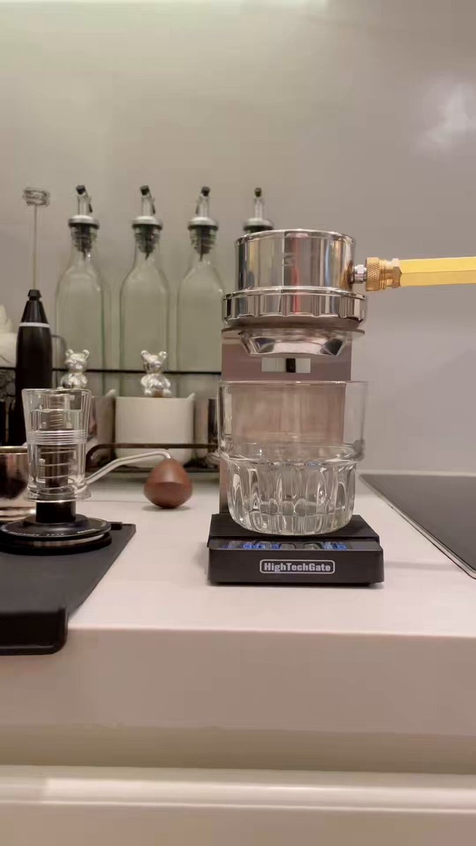 The image features a kitchen counter with various items, including a glass measuring cup sitting on top of a scale that displays "1g Quack!". The scale appears to be used for measuring ingredients or beverages like coffee. There are also several bottles placed around the countertop, possibly containing condiments or other kitchen essentials. A cup is present in the background as well.