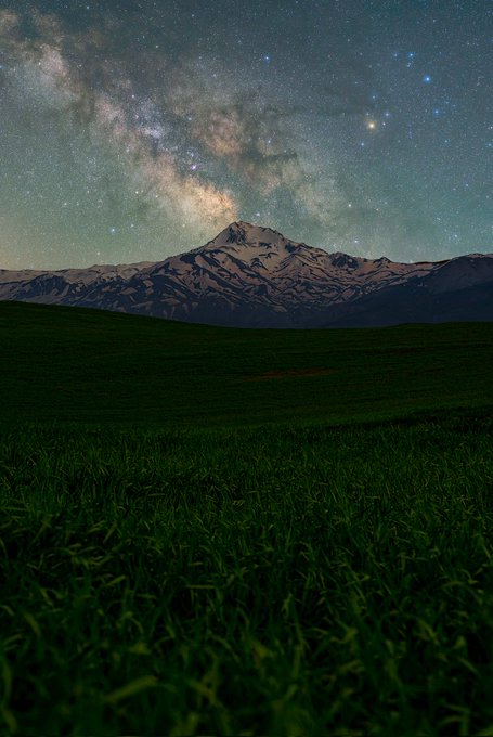The image features a beautiful view of a green field with tall grass, under a starry night sky. The stars are scattered across the sky, creating a captivating backdrop for the scene below. In the distance, there's a mountain range that adds depth and grandeur to the landscape. The photo is taken from a low angle, which emphasizes the height of the mountains and the vastness of the field.