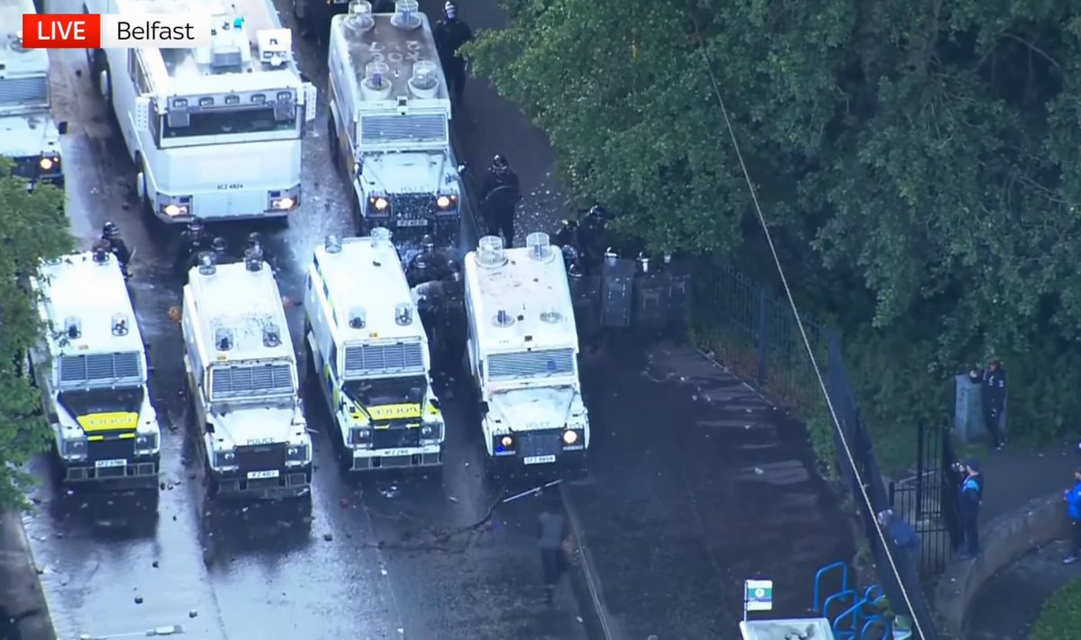 The image shows a group of police vehicles parked next to each other, with some people standing nearby. There are at least six white police vans visible in the scene. The presence of multiple police cars suggests that there might be an ongoing operation or event requiring additional law enforcement support.