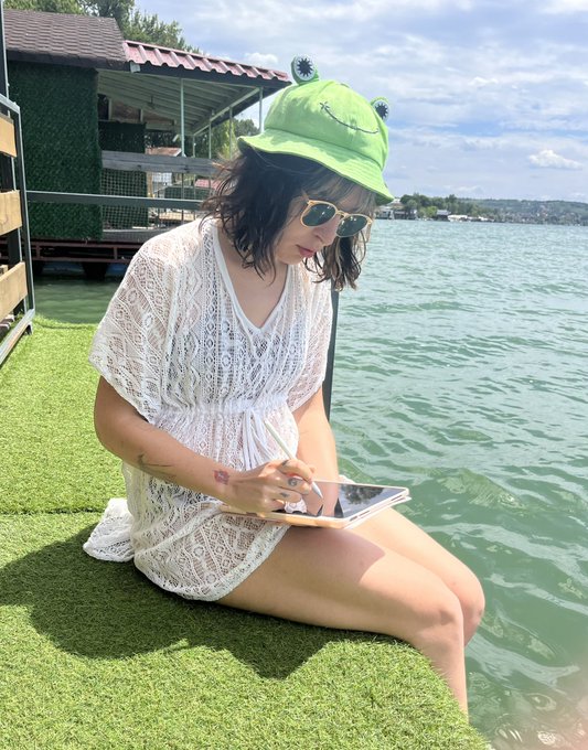 A woman in a white lace dress, green frog-themed bucket hat, and sunglasses sits on artificial grass by the water, writing on a notepad with a stylus.