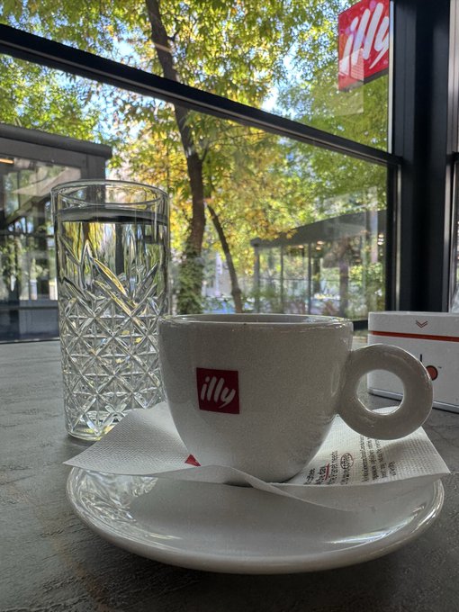 An illy espresso cup and a glass rest on a saucer by a window, with trees and an illy sign visible outside.