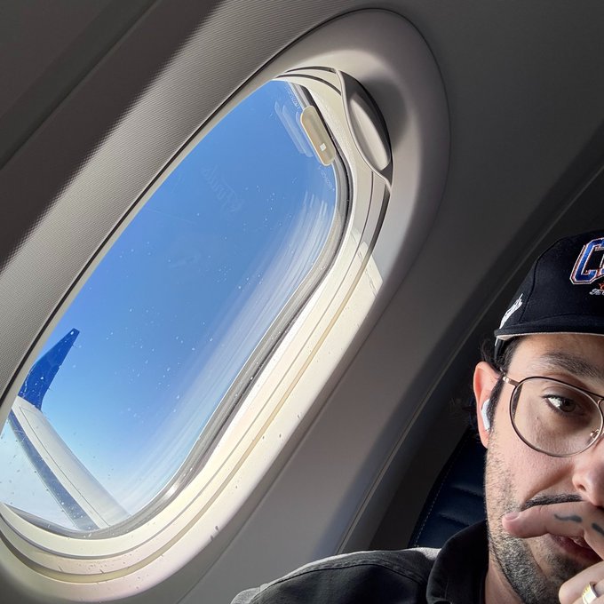 A selfie of a man with glasses and a dark cap with a 'C' logo, taken inside an airplane next to the window showing the sky and wing.