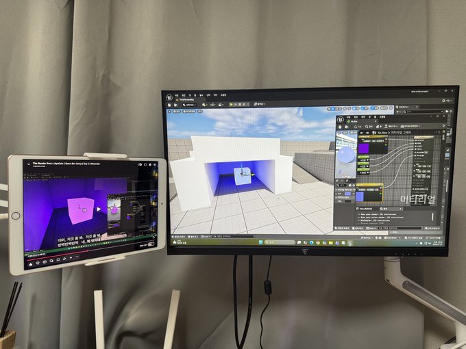 A dual-monitor setup displaying a 3D modeling/rendering software (Unreal Engine) with a tablet on the left mirroring a stylized cube scene, set against a gray curtain backdrop.