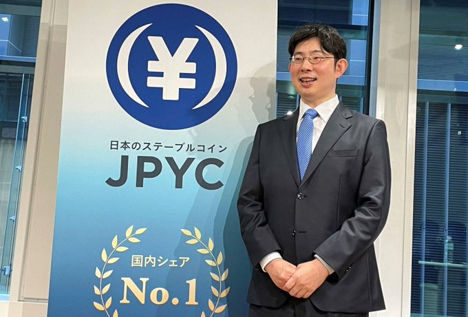 A man in a dark suit and blue tie stands smiling beside a JPYC promotional banner featuring a yen symbol logo and the text "日本のステーブルコイン JPYC" and "国内シェア No.1," visually connecting to news of Lawson's upcoming pilot of the yen-backed JPYC stablecoin for payments.