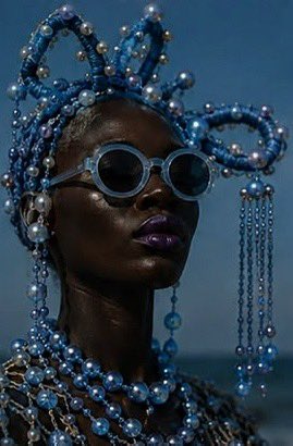 A portrait of a woman wearing an elaborate blue beaded headpiece, round blue sunglasses, and matching beaded jewelry against a clear sky.