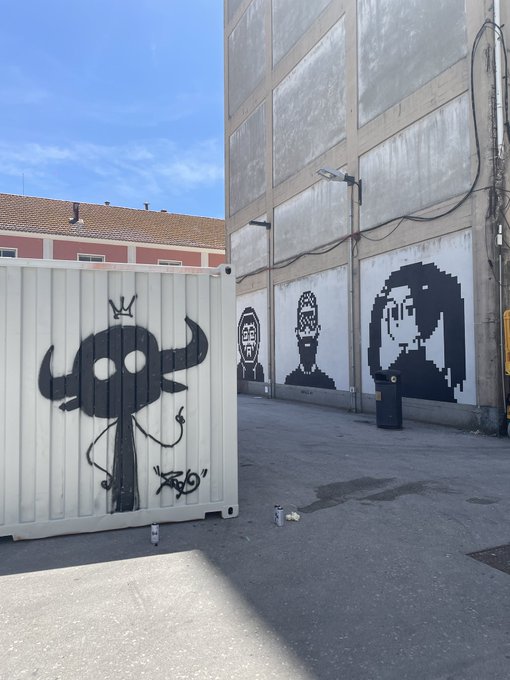 The image features a white building with black graffiti art, including a large skull and two cartoon faces painted on its side. The building also has a sign that reads "Gm. Something coming soon and @normiesART know it". This suggests that there might be an upcoming event or gathering related to the graffiti art displayed on the building.

The image is taken from a low angle, which emphasizes the height of the building and the artwork. The presence of the graffiti adds a sense of urbanity and artistic expression to the scene.