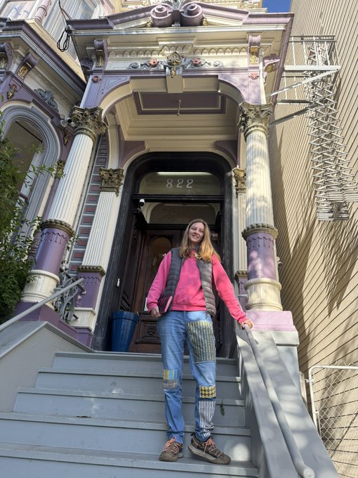 A young woman stands on the steps of a beautifully ornate Victorian-style building marked "822," wearing colorful patchwork pants and a pink top, capturing the moment as she introduces herself from San Francisco after recently arriving from Tel Aviv.
