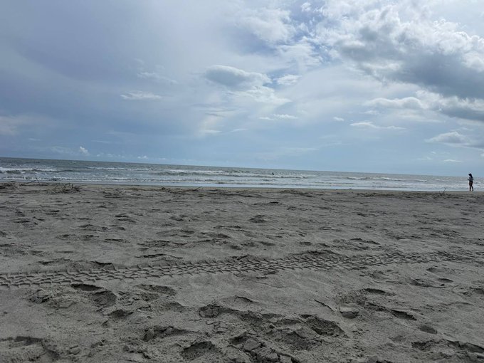 A scenic view of a sandy beach under a cloudy sky with tire tracks and footprints in the sand, with a person standing by the water.