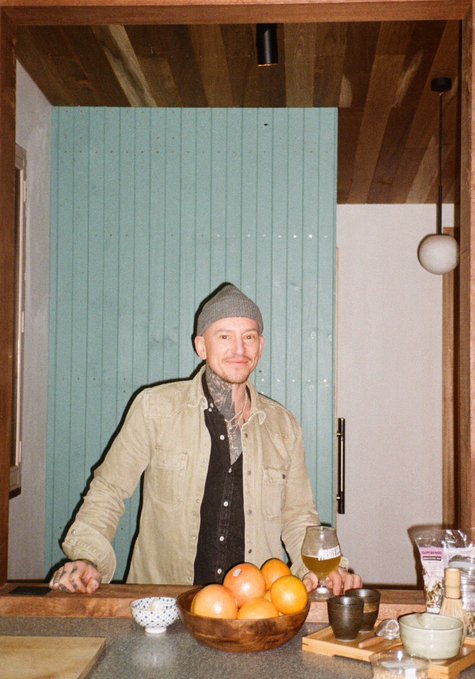 A man wearing a beanie and tan jacket stands behind a kitchen counter with a bowl of oranges and a glass of beer, in what appears to be a casual home setting. The image accompanies a post expressing pride in being a "Bored Ape" member of an online club/community, suggesting the man identifies with or represents this NFT community.