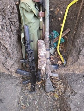 A Ukrainian soldier's gear, including an AK-74 rifle with "XantaresPeek" written on it, rests against the dirt wall of a trench or foxhole.