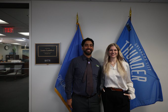 A man and woman stand smiling in front of a 'James Lankford, Oklahoma' Senate plaque and two blue flags reading 'OKLAHOMA CITY THUNDER'.