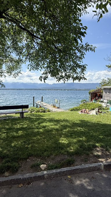 The image shows a serene scene of a lake with a dock, where people are enjoying their time relaxing by the water. There's a bench near the dock, providing a place for visitors to sit and take in the view. The calm waters of the lake reflect the clear blue sky above, creating a peaceful atmosphere. In the background, there is a house visible on one side of the lake, adding to the picturesque setting.