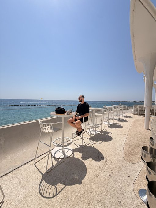 A man in sunglasses and a black t-shirt sits alone at a high-top table along a sunlit seaside terrace overlooking the Mediterranean, embodying the post's narrative of trading a corporate job for a European coastal lifestyle. The minimalist white barstools and pristine ocean view visually reinforce the message that manifesting a dramatically better life is achievable.
