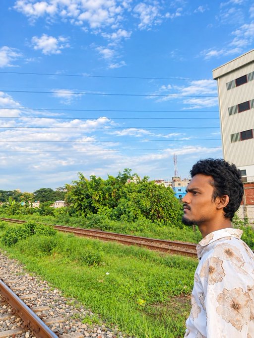 A young man with dark curly hair wearing a floral shirt stands on the right side of the frame, looking out at railway tracks surrounded by green vegetation and buildings under a bright blue sky.