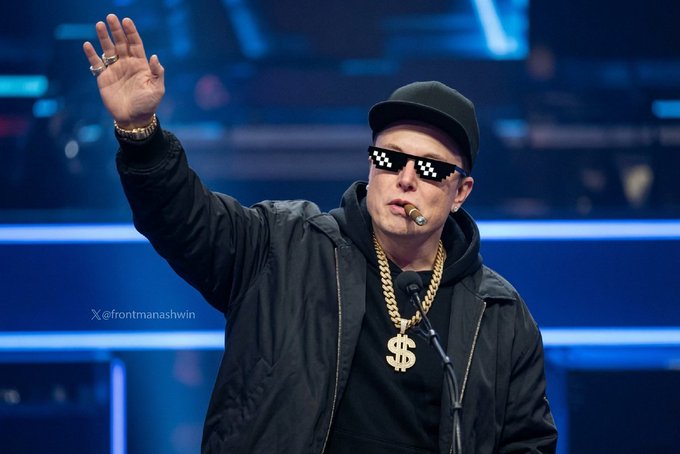 A man wearing pixelated sunglasses, a black cap, and gold chains with a dollar sign pendant, waving at what appears to be a conference or event.