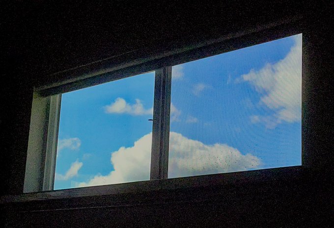 A view through a dark window frame showing a bright blue sky with scattered white clouds, with visible dust particles on the glass.