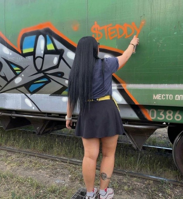 A young woman with long black hair, wearing a black skirt and gray top, is seen from behind spray-painting an orange 'STEDDY' tag onto a green freight train car.