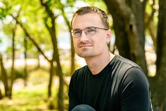 A man with glasses and a beard wearing a black t-shirt, sitting outdoors with a blurred green background of trees.