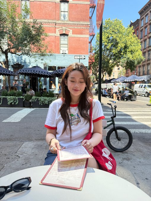 A young woman with long brown hair sits at an outdoor cafe table reading a menu; she wears a white ringer t-shirt with red trim and has sunglasses resting on the table.