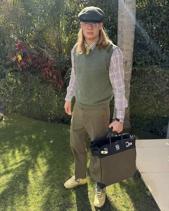 A young man with long hair, glasses, and a flat cap poses in a garden wearing a layered outfit of a checkered shirt under a green sweater vest with olive trousers, holding a large Hermès Birkin-style bag.