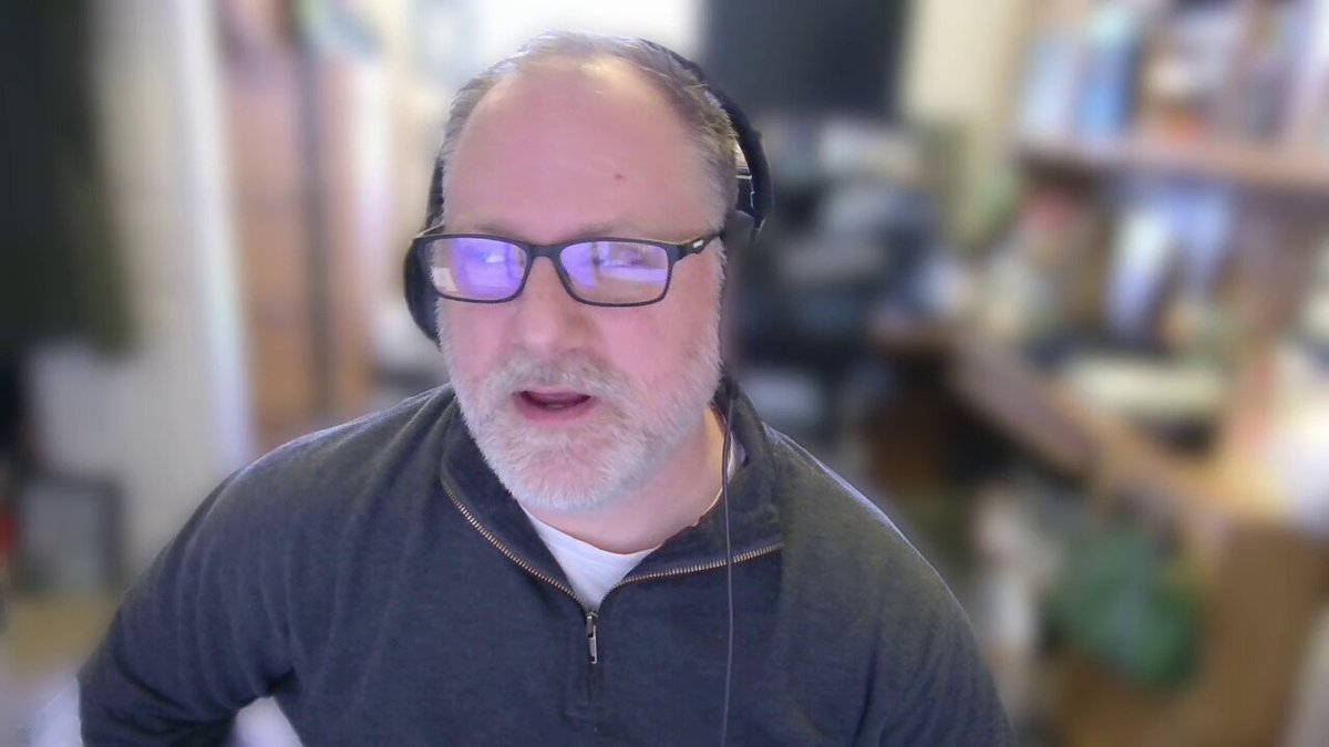 The image features a man wearing glasses, possibly an older gentleman with gray hair, who appears to be talking into a microphone or headset. The background of the image shows a bookshelf filled with numerous books, suggesting that he might be in a library or a room dedicated to reading and learning.