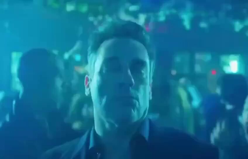 The image shows a man with short hair wearing a black shirt, standing in front of a blue background that appears to be a nightclub or bar setting. The man has his eyes closed, suggesting he might be meditating or taking a break from dancing. There are other people visible in the scene as well, indicating that this is likely an indoor social event with music and dancing.