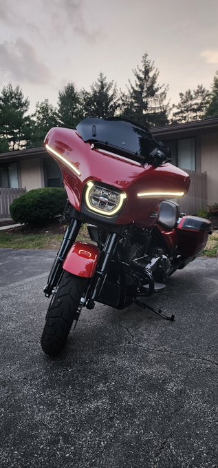 A sleek red touring motorcycle, likely a Harley-Davidson Road Glide, stands parked in a driveway at dusk with its LED headlights and signature front fairing glowing prominently. The image perfectly complements the caption's sentiment of admiration, showcasing the bike as a prized possession rivaling Bitcoin in personal significance.