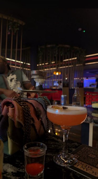 Cocktail at a dimly lit bar - an orange/peach colored drink in a coupe glass, alongside a shot glass with red liquid, with a person in the background.