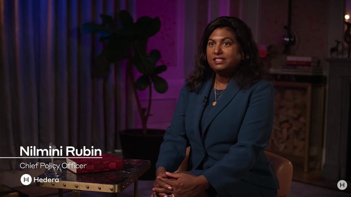 The image features a woman sitting at a desk, wearing a blue suit jacket. The background of the image shows a bookshelf filled with various books. There is also a potted plant visible behind her. The caption for the image reads "America's biggest banks are reportedly building a tokenised deposit network could be in the conversation @hedera 's head of policy is says here they are already speaking with policymakers and big banks!".