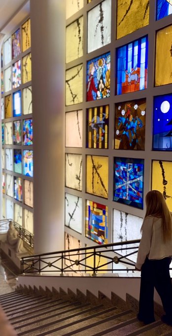 A woman walks up a staircase next to a tall, curved wall composed of numerous illuminated stained glass panels depicting religious imagery in blue, yellow, and white tones.
